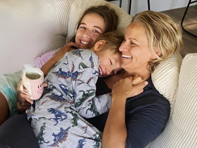 A smiling woman cuddles a young boy in dinosaur pajamas and a smiling girl on a cozy couch. The woman holds a mug. All three look happy and relaxed, enjoying a close, affectionate moment together.