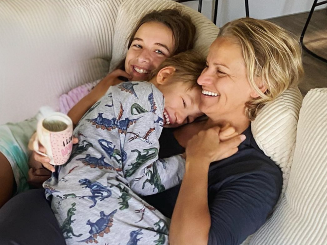 A smiling woman cuddles a young boy in dinosaur pajamas and a smiling girl on a cozy couch. The woman holds a mug. All three look happy and relaxed, enjoying a close, affectionate moment together.