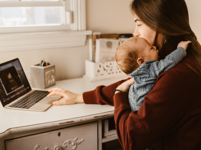 A woman holds a baby while working on a laptop at a white desk near a window, multitasking between childcare and work in a cozy home environment.