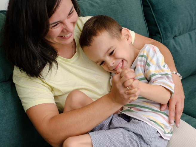 A woman and a young boy sit on a green couch, smiling and laughing as they hug and play together. The boy wears a striped shirt and shorts, and the woman is dressed in a yellow top.