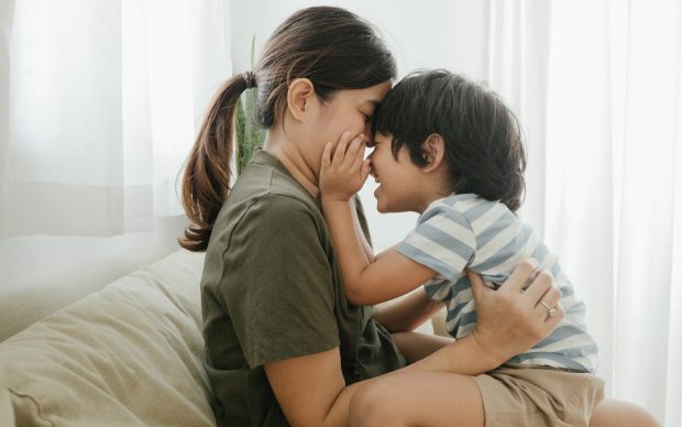 A woman and a young child sit face-to-face on a couch, smiling and touching noses affectionately, with sunlight streaming through white curtains in the background.