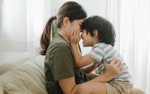 A woman and a young child sit face-to-face on a couch, smiling and touching noses affectionately, with sunlight streaming through white curtains in the background.