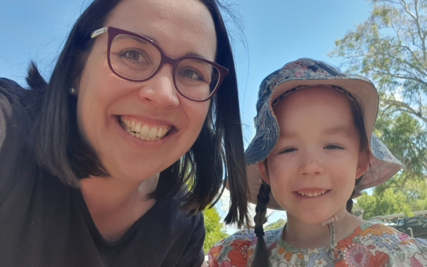 A woman with glasses and a young girl wearing a floral hat and dress smile outdoors on a sunny day, with trees and a bright blue sky in the background.