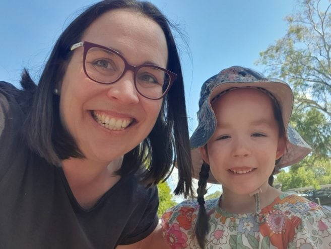 A woman with glasses and a young girl wearing a floral hat and dress smile outdoors on a sunny day, with trees and a bright blue sky in the background.
