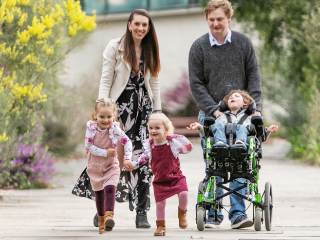 A smiling family walks outside. Two young girls holding hands run in front, while a woman and a man walk behind them. The man pushes a boy in a wheelchair. They are surrounded by blooming plants.
