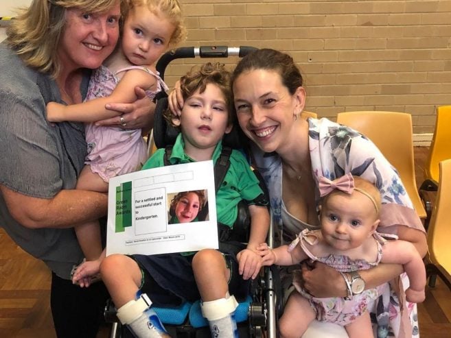 A smiling group poses indoors: an older woman holds a young girl, a boy sits in a wheelchair holding a sign, a woman crouches beside them, and a baby in a bow headband sits on her lap.
