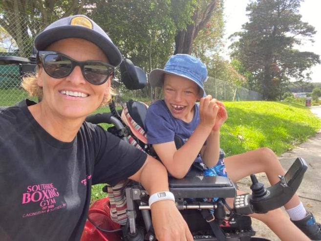 A smiling woman wearing sunglasses and a cap takes a selfie with a joyful boy in a motorized wheelchair outdoors on a sunny day, with trees and grass in the background.