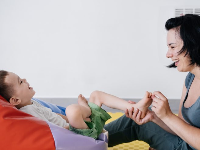 A woman smiles and tickles the bare foot of a young child who is laughing while lying on a colorful bean bag. They are playing together on a soft, yellow play mat indoors.