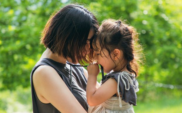 A woman and a young girl stand close together outdoors, touching foreheads and sharing an affectionate moment, surrounded by greenery and sunlight.