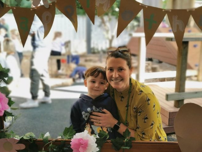 A smiling woman in a yellow dress and a young boy stand together by a decorated garden frame with pink flowers and bunting, outdoors, with children playing in the blurred background.