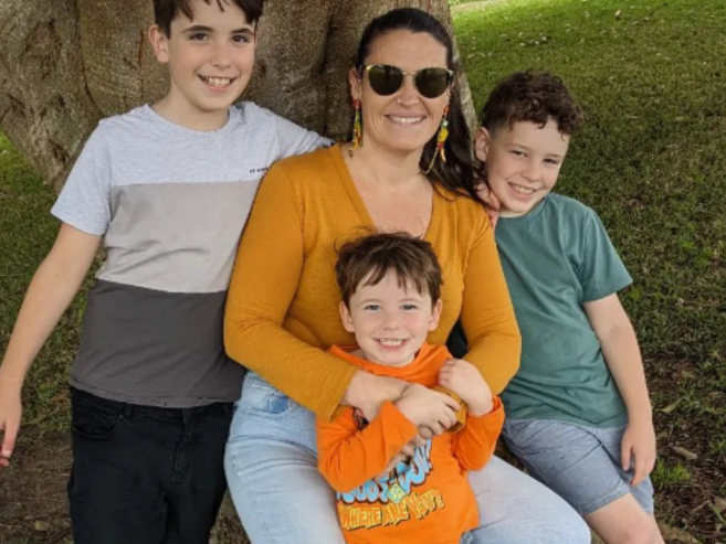 A woman wearing sunglasses and an orange shirt sits under a tree, smiling with three young boys. The boys are hugging her, and they all look happy. Grass and tree roots are visible in the background.