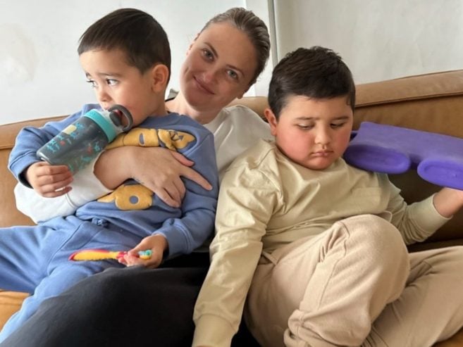 A woman sits on a brown couch, smiling and hugging two young boys. One boy holds a water bottle and a toy, while the other boy rests, holding a purple foam object.