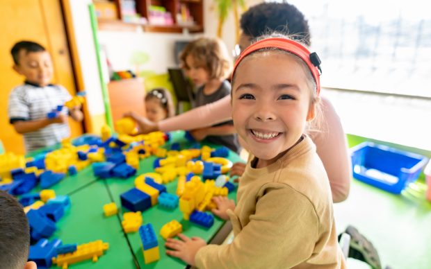 Smiling young girl at a table with colorful plastic building blocks, surrounded by other children playing and building in a bright, cheerful classroom.