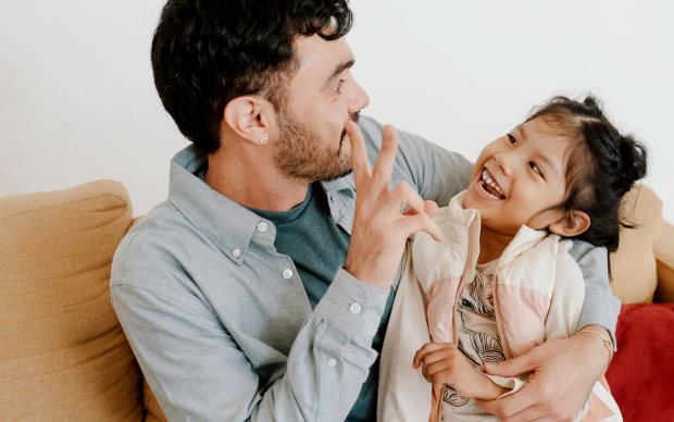 A man and a young girl sit on a couch, smiling and laughing together. The man gently holds the girl with one arm while playfully gesturing with his hand near her face. They both appear happy and relaxed.