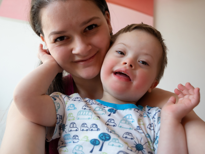 A smiling woman hugs a cheerful young boy with Down syndrome. The boy is wearing a shirt with colorful cars and trees, and both are looking happily at the camera in a bright indoor space.