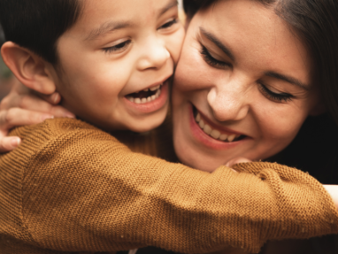 A young child and an adult woman share a joyful embrace, both smiling warmly. The child wears a brown sweater and their faces are close together, expressing happiness and affection.