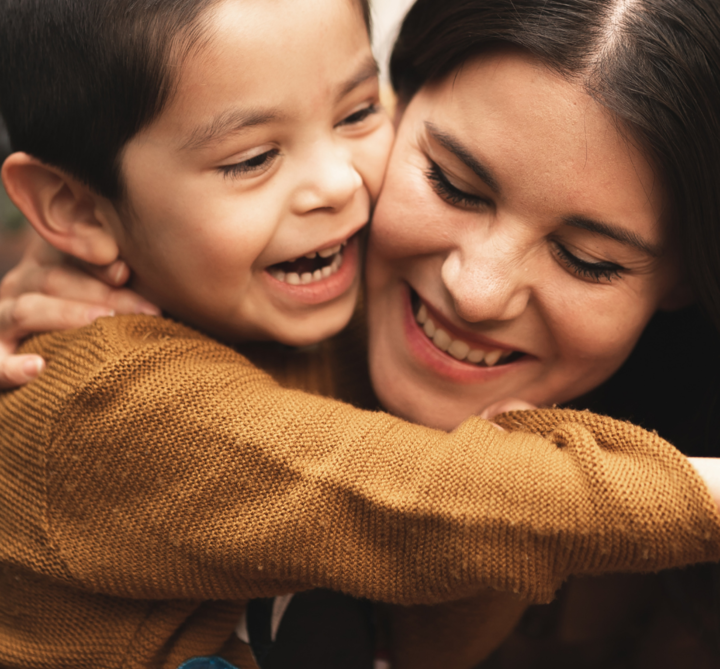 A young child and an adult woman share a joyful embrace, both smiling warmly. The child wears a brown sweater and their faces are close together, expressing happiness and affection.
