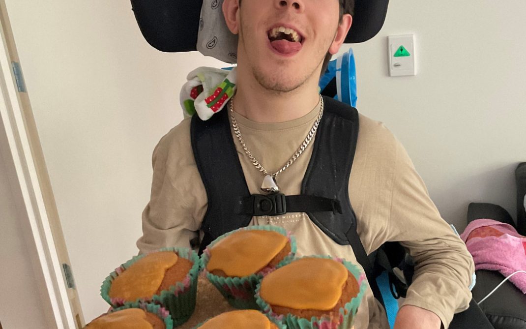 A young man in a wheelchair is smiling and holding a round tray with five cupcakes topped with caramel icing. He is wearing glasses, a beige shirt, and a silver chain necklace in a home setting.