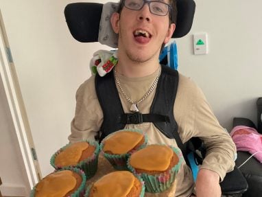 A young man in a wheelchair is smiling and holding a round tray with five cupcakes topped with caramel icing. He is wearing glasses, a beige shirt, and a silver chain necklace in a home setting.