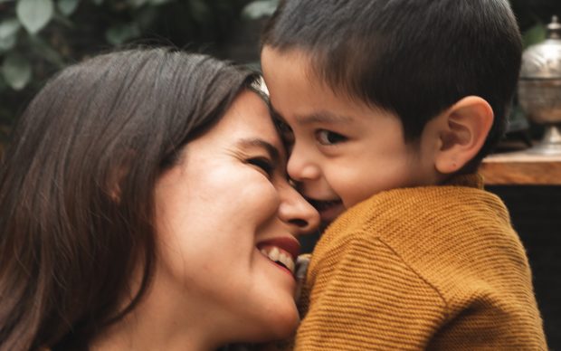 A woman and a young boy, both wearing brown jackets, share a joyful and affectionate moment, smiling closely with noses touching against a leafy green background.