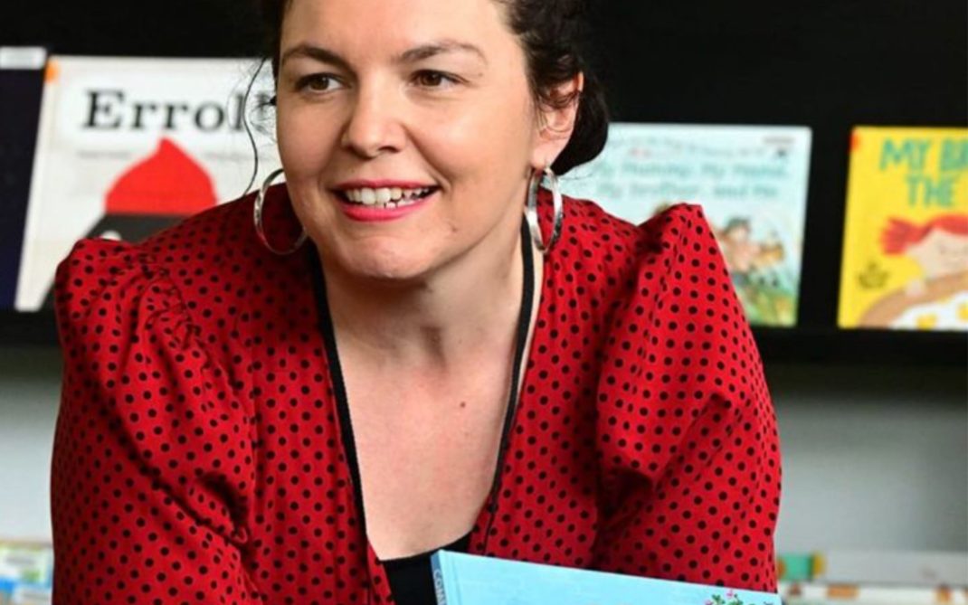 A woman with dark curly hair in a red polka-dot blouse is smiling and holding a childrens book titled Come Over to My House, with books displayed on a shelf in the background.