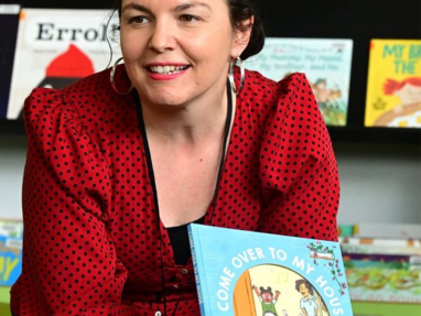 A woman with dark curly hair in a red polka-dot blouse is smiling and holding a childrens book titled Come Over to My House, with books displayed on a shelf in the background.