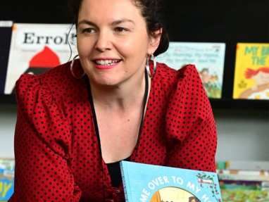A woman with dark curly hair in a red polka-dot blouse is smiling and holding a childrens book titled Come Over to My House, with books displayed on a shelf in the background.
