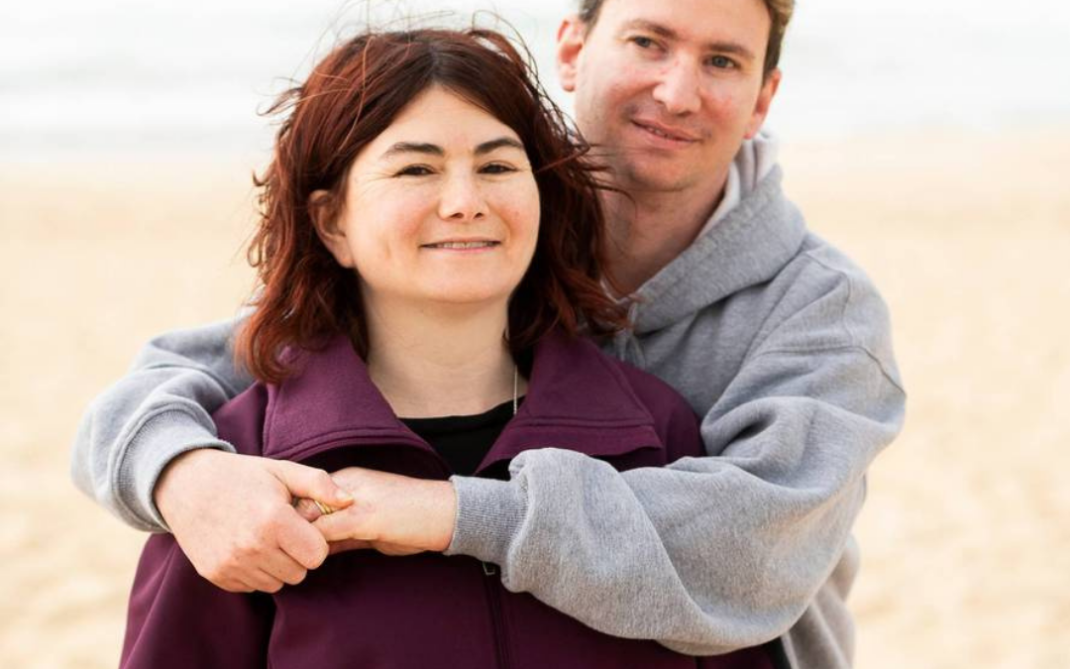A man stands behind a woman on a beach, smiling and hugging her from behind. The woman wears a purple jacket, and the man wears a gray hoodie. The ocean and sand are visible in the background.
