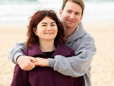 A man stands behind a woman on a beach, smiling and hugging her from behind. The woman wears a purple jacket, and the man wears a gray hoodie. The ocean and sand are visible in the background.