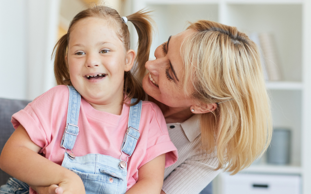 A smiling young girl with Down syndrome wearing denim overalls and a pink shirt sits on a couch while a woman with blonde hair hugs her from behind, both appearing happy and playful.