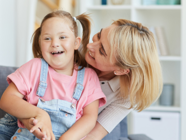 A smiling young girl with Down syndrome wearing denim overalls and a pink shirt sits on a couch while a woman with blonde hair hugs her from behind, both appearing happy and playful.