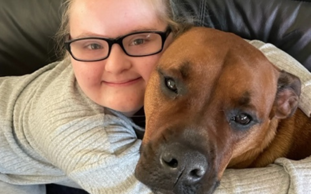 A smiling person wearing glasses and a grey top hugs a large brown dog on a black leather couch. Both look content and relaxed.