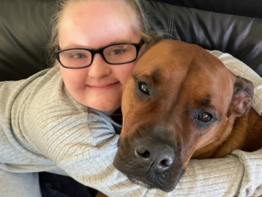 A smiling person wearing glasses and a grey top hugs a large brown dog on a black leather couch. Both look content and relaxed.