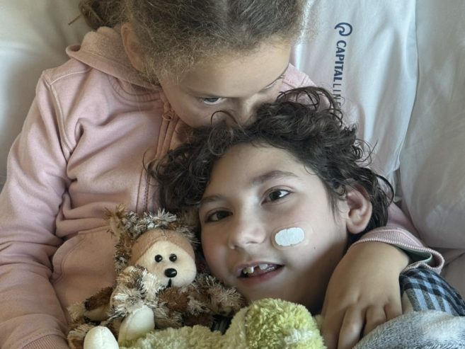 A child in a hospital bed, wearing a nasal bandage and hospital bracelet, cuddles a stuffed animal while being embraced by an older child. Both appear calm and affectionate, wrapped in a cozy blanket.