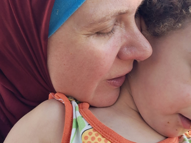 A woman in a maroon hijab lovingly hugs a young child outdoors. Sunlight filters through green foliage behind them as the woman gently presses her face to the child’s, both eyes closed in a tender moment.