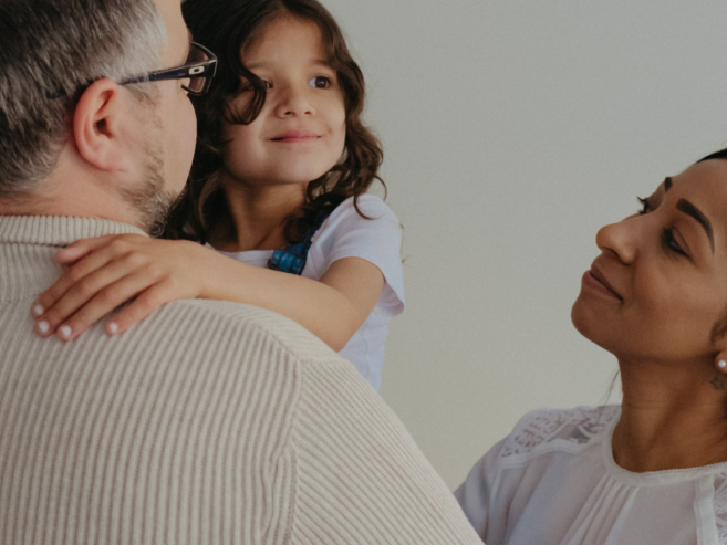 A man with glasses holds a young girl, while a woman smiles at them. The girl looks content, and the family stands together against a plain background.