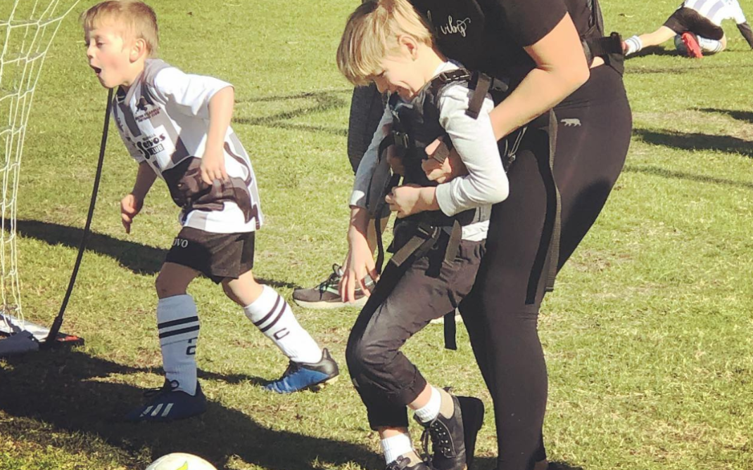A woman assists a young boy with mobility needs in kicking a soccer ball on a grassy field, while another child in a soccer uniform runs nearby. Other children play in the background.