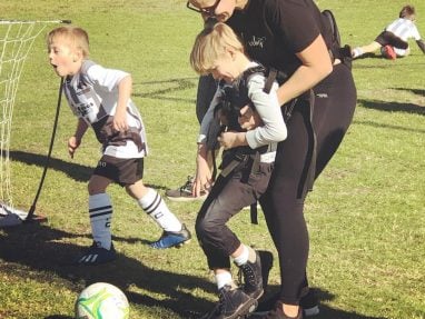 A woman assists a young boy with mobility needs in kicking a soccer ball on a grassy field, while another child in a soccer uniform runs nearby. Other children play in the background.