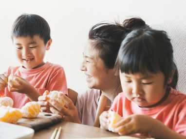 A smiling woman sits at a table with two children, all wearing pink shirts, as they peel oranges together. The children focus on peeling while the woman looks at them happily.