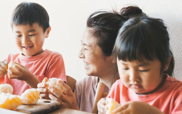 A smiling woman sits at a table with two children, all wearing pink shirts, as they peel oranges together. The children focus on peeling while the woman looks at them happily.