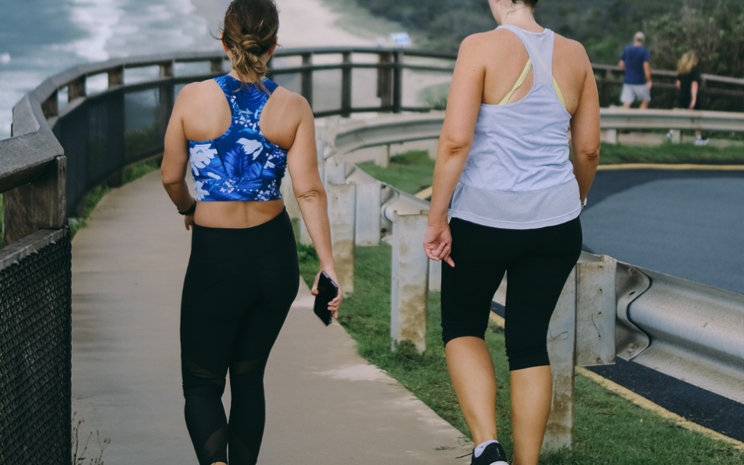 Two women in athletic wear walk on a paved coastal path with a scenic ocean view to their left. One woman wears a blue floral sports bra; the other wears a white tank top. Both are walking away from the camera.