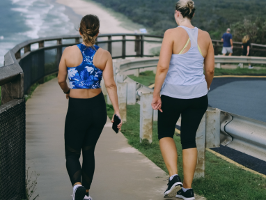 Two women in athletic wear walk on a paved coastal path with a scenic ocean view to their left. One woman wears a blue floral sports bra; the other wears a white tank top. Both are walking away from the camera.