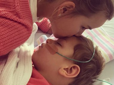 A woman in a pink sweater gently kisses the forehead of a young child lying in bed with a nasal oxygen tube and medical tape on their face, wrapped in a white blanket.