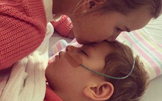 A woman in a pink sweater gently kisses the forehead of a young child lying in bed with a nasal oxygen tube and medical tape on their face, wrapped in a white blanket.