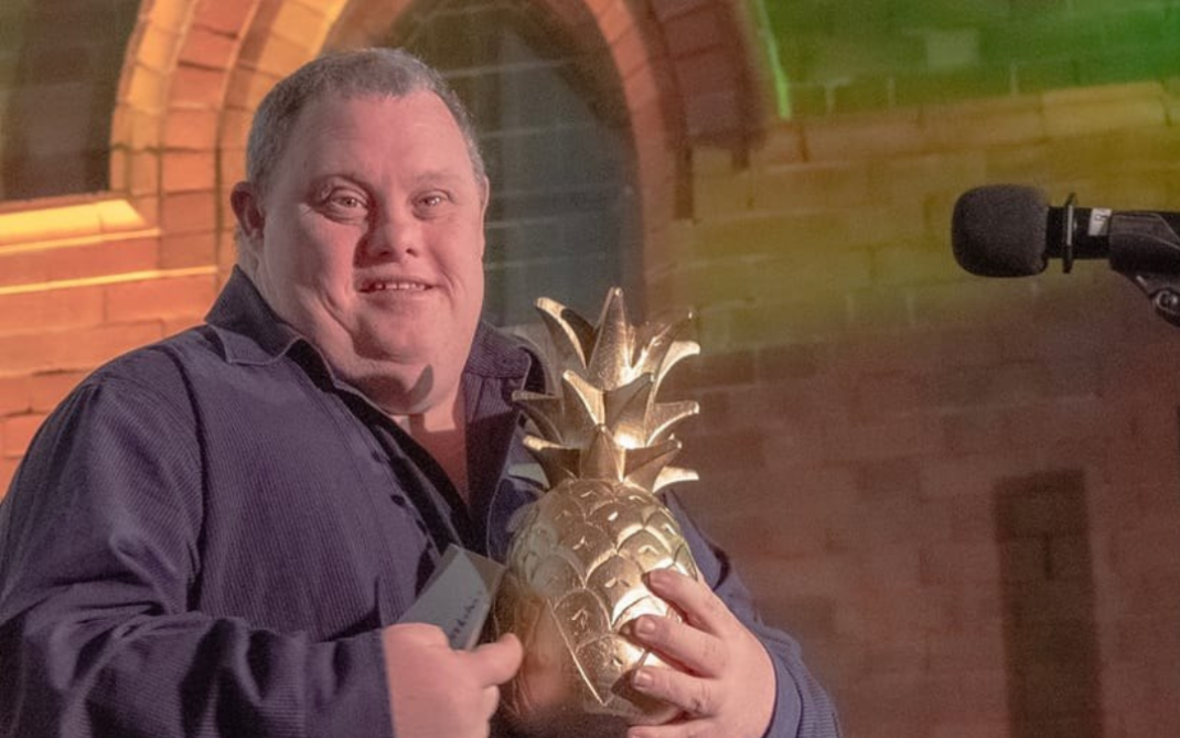 A smiling man wearing a dark shirt holds a large golden pineapple-shaped trophy while standing beside a microphone, with a brick wall and arched window in the background.