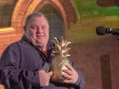 A smiling man wearing a dark shirt holds a large golden pineapple-shaped trophy while standing beside a microphone, with a brick wall and arched window in the background.