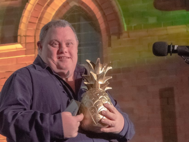 A smiling man wearing a dark shirt holds a large golden pineapple-shaped trophy while standing beside a microphone, with a brick wall and arched window in the background.