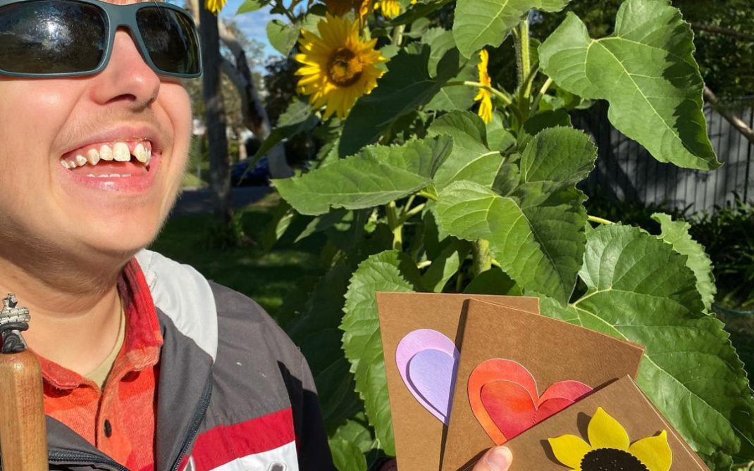 A smiling person wearing sunglasses holds three handmade cards with heart and sunflower designs, standing beside tall blooming sunflowers in bright sunlight.