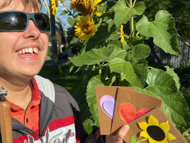 A smiling person wearing sunglasses holds three handmade cards with heart and sunflower designs, standing beside tall blooming sunflowers in bright sunlight.
