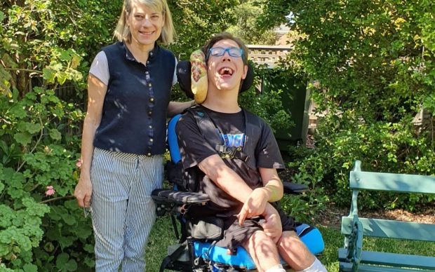 A smiling woman stands next to a young man in a motorized wheelchair with a therapy device on his head. They are outdoors in a garden with green plants, trees, and a green bench nearby.